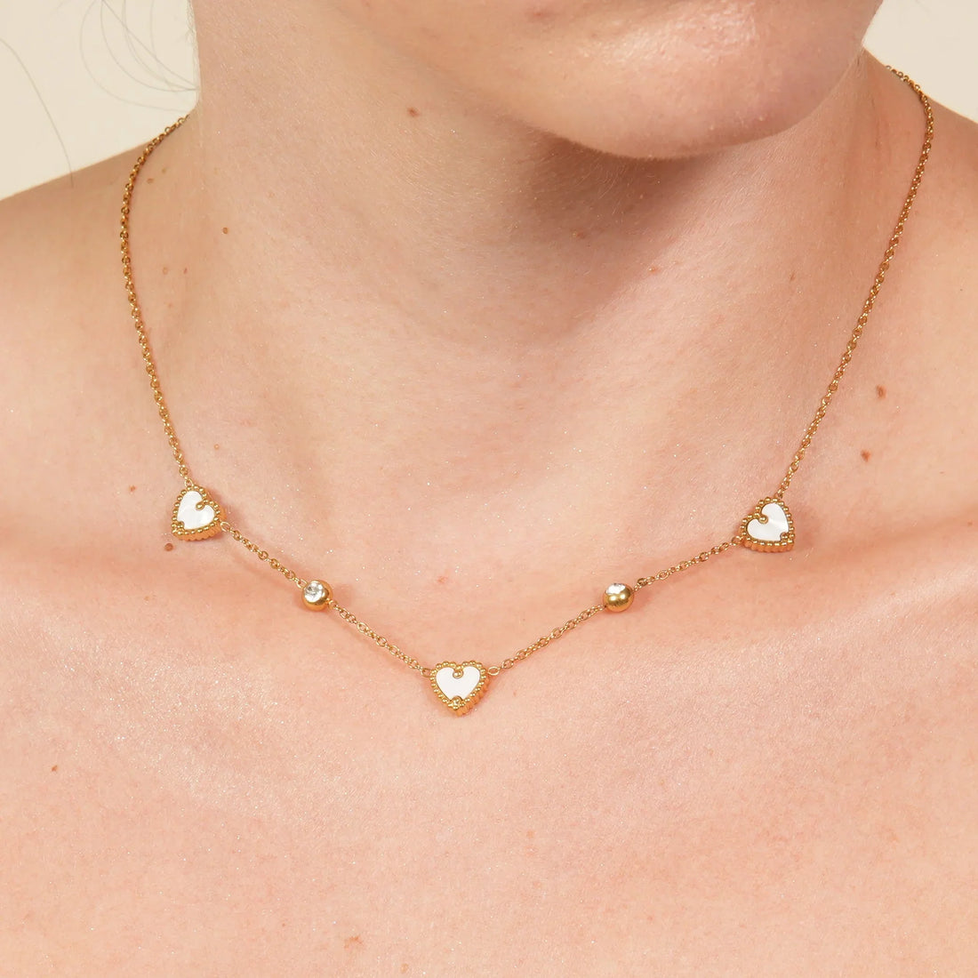 Gold chain necklace with small white heart charms and bead accents worn on a woman's neck, minimalist romantic jewelry design