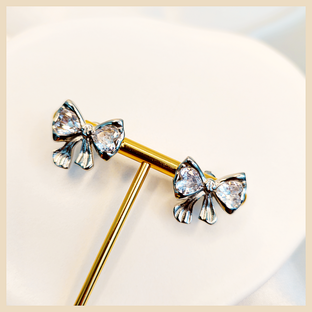 Cadi bow huggie earrings in silver with delicate bow stud detailing, perfect for women who love romantic jewellery that feels polished and gift-worthy.
