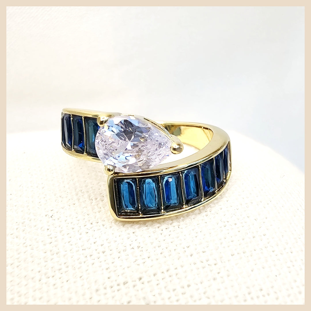 Gold ring with a large clear gemstone and smaller blue gems on a textured beige background